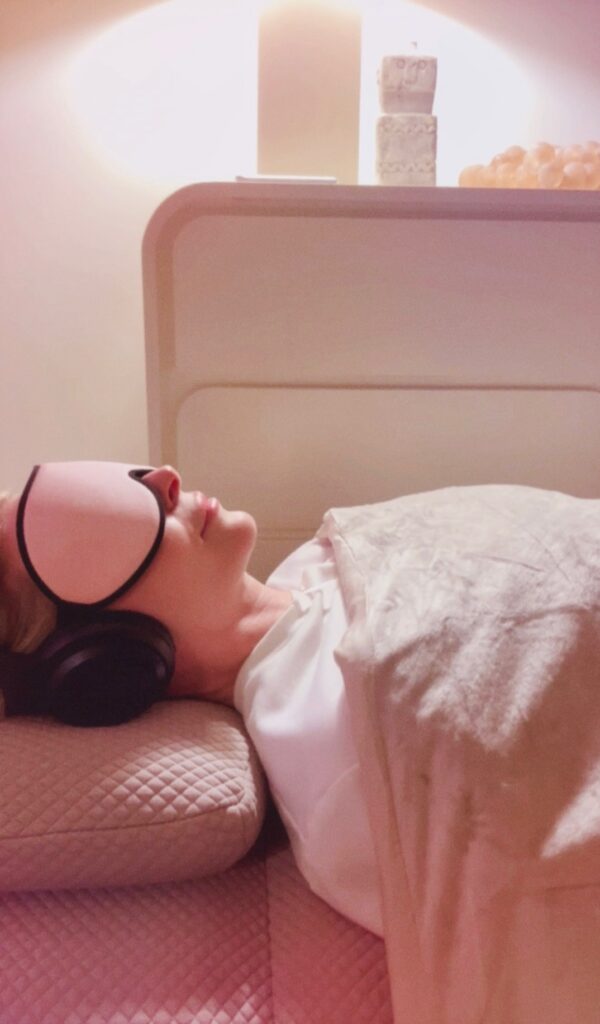 Soundbed therapy session with a woman lying under a blanket wearing headphones and an eye mask in a dim room.