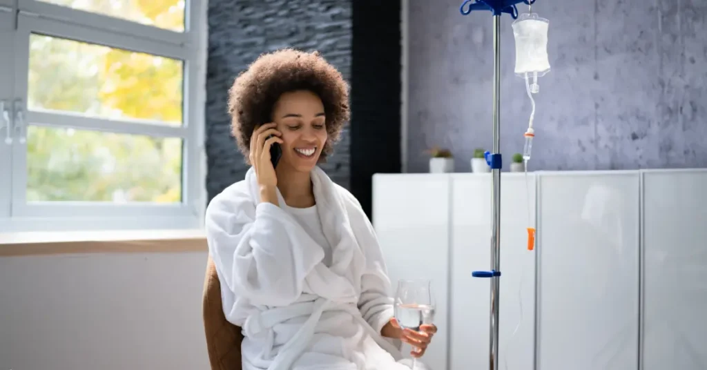 Ketamine therapy clinic patient receiving IV while talking on phone and holding a glass of water.