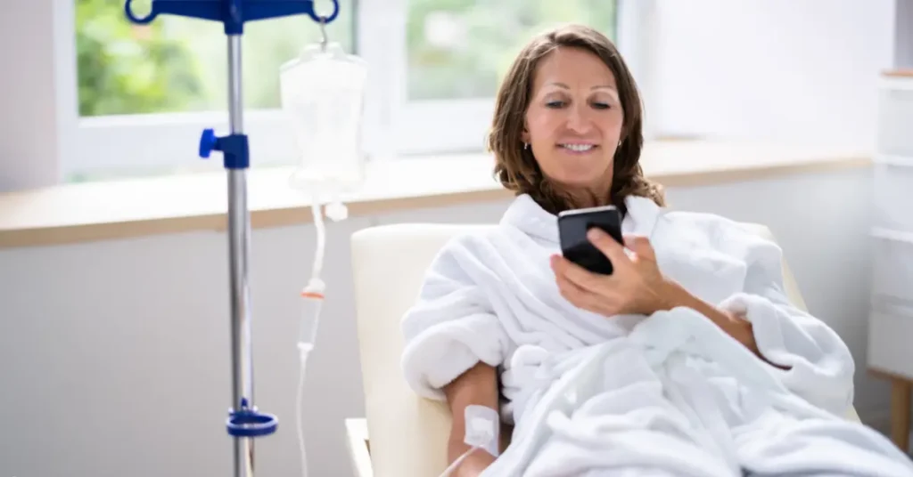 Psychedelic-assisted therapy image of woman relaxing in chair with IV and smiling while using smartphone.
